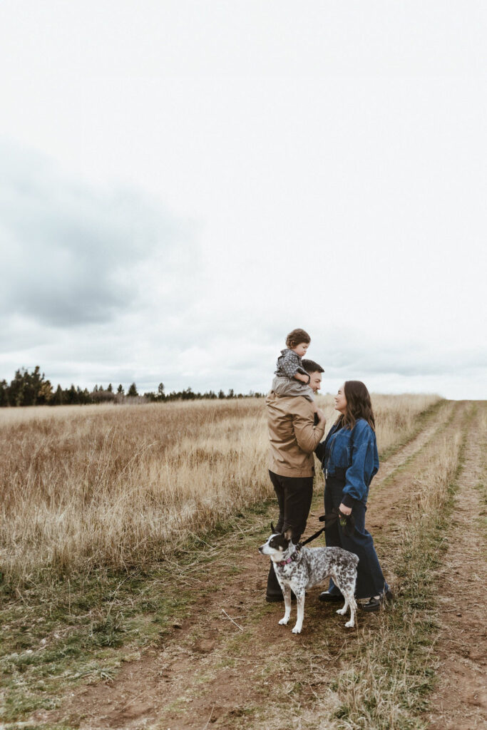 A faraway photo of this family of 3 with their family dog, where mom and dad look at each other in a beautiful wheat field in a Spokane family portrait session with Sophie Grace Photography