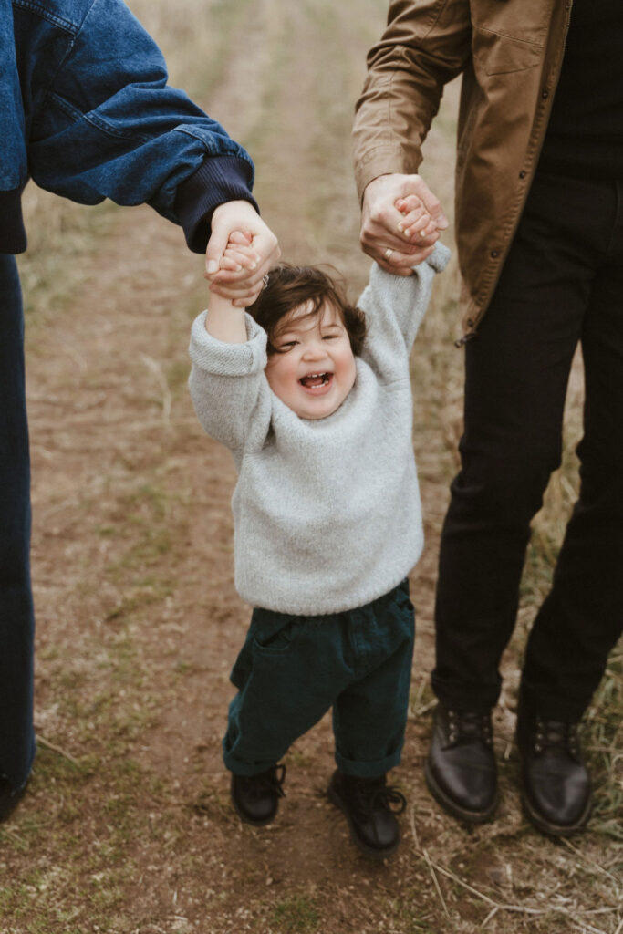 Little boy laughing walking with mom and dad holding his hands on both sides during a family session in Spokane, WA. Sophie Grace Photography