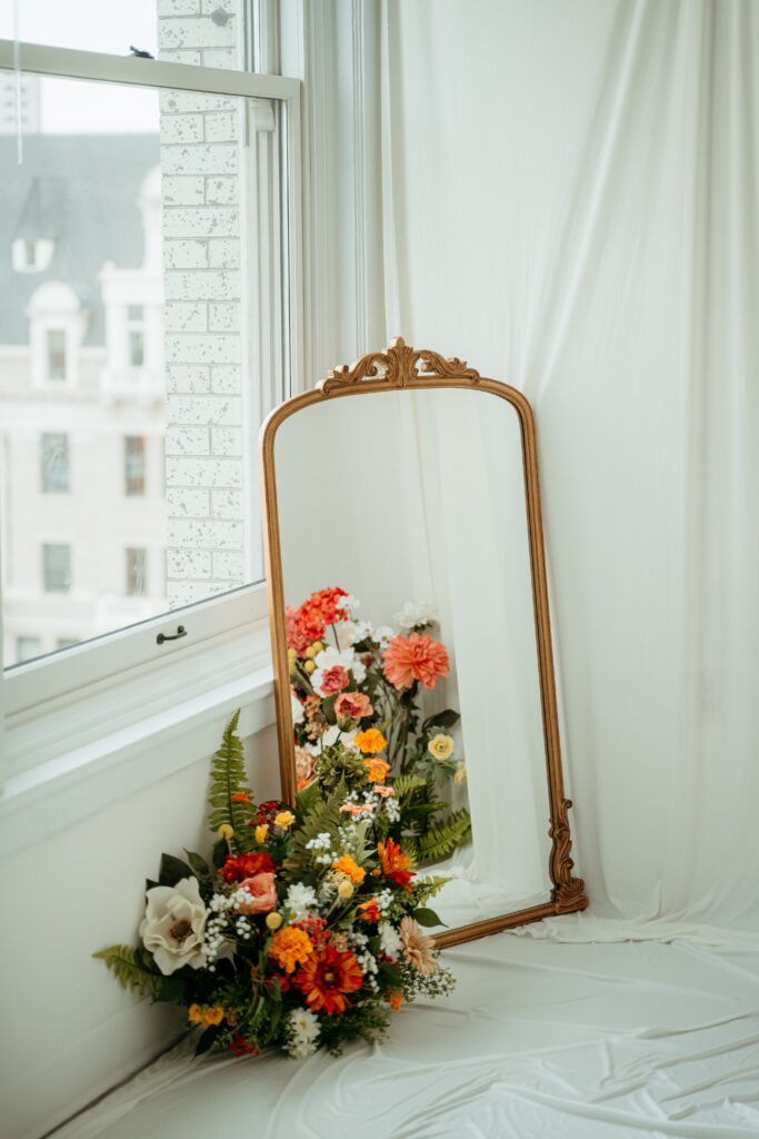 Spring studio motherhood photography setup in Spokane WA featuring an antique gold mirror, soft white draping, and colorful spring florals by a window.